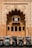 In front of an intricately designed historical building with an ornate archway featuring carved fish motifs, several auto rickshaws are parked in a row. The building showcases traditional architectural elements with arched windows and detailed stonework. The scene suggests a blend of cultural heritage and modern transportation.