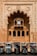 In front of an intricately designed historical building with an ornate archway featuring carved fish motifs, several auto rickshaws are parked in a row. The building showcases traditional architectural elements with arched windows and detailed stonework. The scene suggests a blend of cultural heritage and modern transportation.