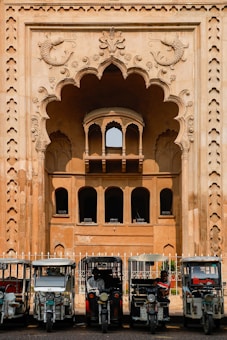 In front of an intricately designed historical building with an ornate archway featuring carved fish motifs, several auto rickshaws are parked in a row. The building showcases traditional architectural elements with arched windows and detailed stonework. The scene suggests a blend of cultural heritage and modern transportation.