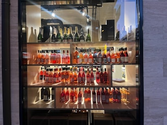 A display shelf filled with various bottles of wine and champagne. The top shelf features champagne bottles, while the middle and lower shelves contain bottles of rosé wine. The setting is well-lit, highlighting the elegant arrangement of the bottles against a neutral background.