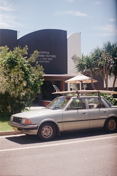 A vintage car is parked on the side of a street with surfboards attached to a roof rack. In the background, a building with signage for McTavish Surfboard Factory and a cafe is partially visible, surrounded by trees and plants.