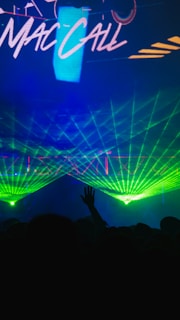 A vibrant display of green laser lights creates a dynamic visual spectacle against a dark background. The silhouette of a person's hand is raised in the foreground, contributing to the vibrant atmosphere of a live performance or event. Bright colors and motion dominate the scene, capturing the essence of an energetic night out.