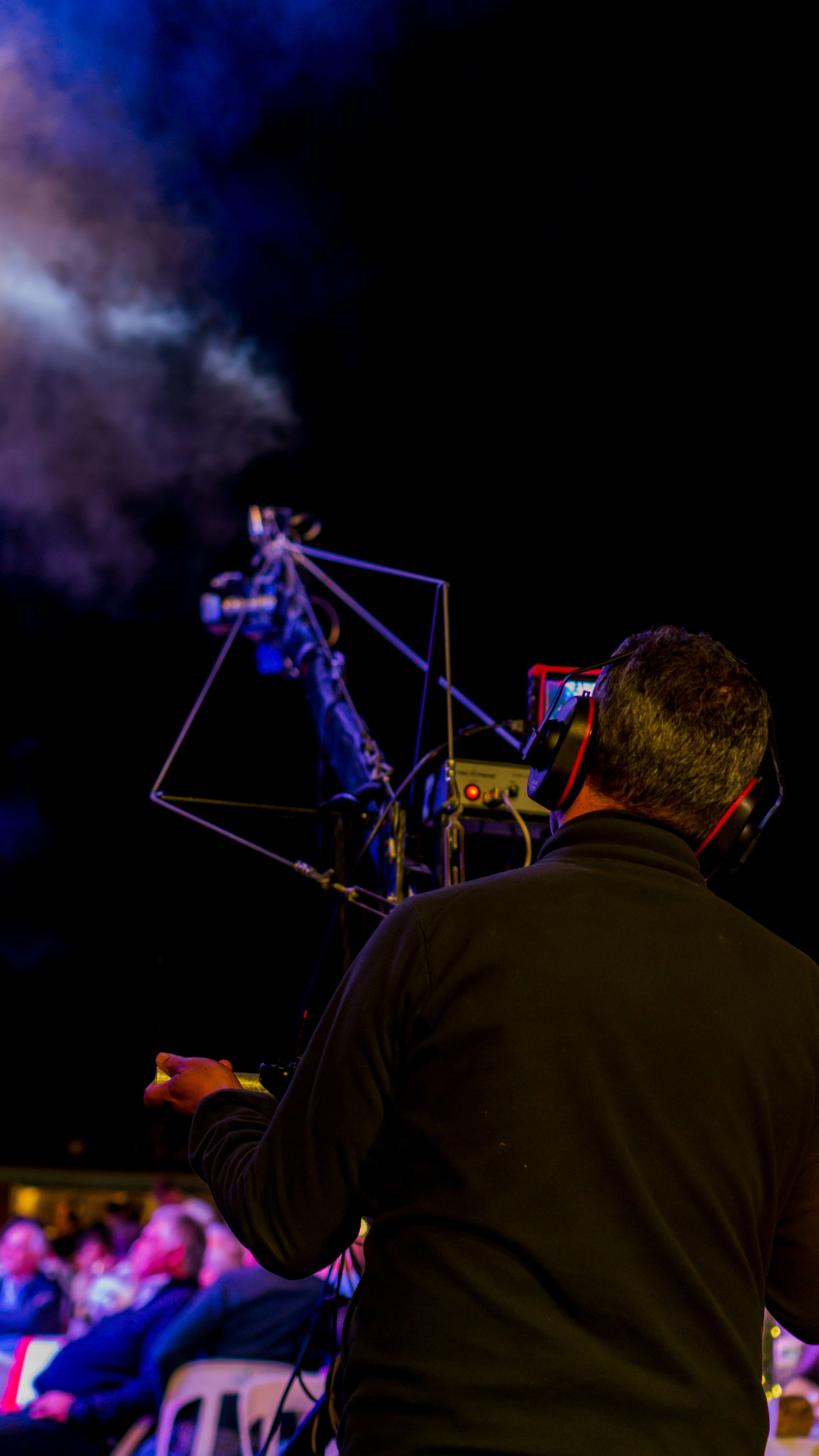 A person operating a camera with a large tripod setup, possibly during an outdoor event. There is a smoky atmosphere visible in the background, and the scene is dimly lit suggesting night time or a low light environment.