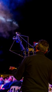 A person operating a camera with a large tripod setup, possibly during an outdoor event. There is a smoky atmosphere visible in the background, and the scene is dimly lit suggesting night time or a low light environment.