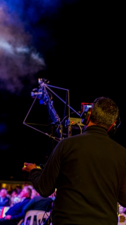 A person operating a camera with a large tripod setup, possibly during an outdoor event. There is a smoky atmosphere visible in the background, and the scene is dimly lit suggesting night time or a low light environment.