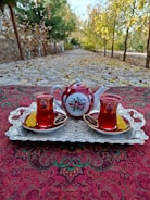A richly decorated red teapot and two matching glass cups filled with tea sit on a vintage silver tray. The tray rests on a patterned tablecloth with intricate designs. Slices of dried citrus and sugar sticks accompany the cups. In the background, a tree-lined path with autumn leaves scattered along the ground adds a rustic charm.