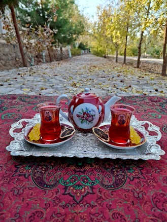 A richly decorated red teapot and two matching glass cups filled with tea sit on a vintage silver tray. The tray rests on a patterned tablecloth with intricate designs. Slices of dried citrus and sugar sticks accompany the cups. In the background, a tree-lined path with autumn leaves scattered along the ground adds a rustic charm.
