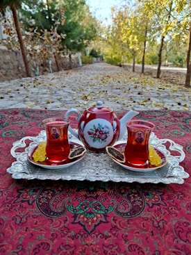 A richly decorated red teapot and two matching glass cups filled with tea sit on a vintage silver tray. The tray rests on a patterned tablecloth with intricate designs. Slices of dried citrus and sugar sticks accompany the cups. In the background, a tree-lined path with autumn leaves scattered along the ground adds a rustic charm.