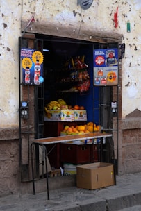 A small street-side shop with an open doorway. Inside, there are stacked boxes of fruit, including bananas and mangoes. Various packaged snack items hang on shelves above the fruit. Advertising posters with colorful text are on both sides of the doorframe. A brown cardboard box is on the ground outside the shop.