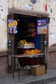 A small street-side shop with an open doorway. Inside, there are stacked boxes of fruit, including bananas and mangoes. Various packaged snack items hang on shelves above the fruit. Advertising posters with colorful text are on both sides of the doorframe. A brown cardboard box is on the ground outside the shop.