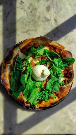 A pizza topped with fresh greens and a large dollop of creamy white cheese sits on a marble surface. The crust is golden brown with slightly charred edges, and the sunlight casts dynamic shadows across the scene.