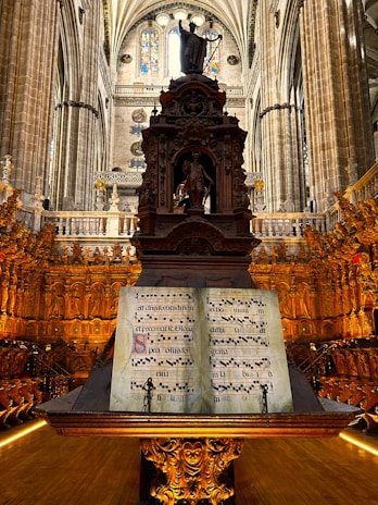 Intricately designed cathedral interior featuring ornate wood carvings and a large music stand with an illuminated manuscript. A statue stands atop the ornate structure amidst towering columns and stained glass windows, creating a majestic and reverent atmosphere.
