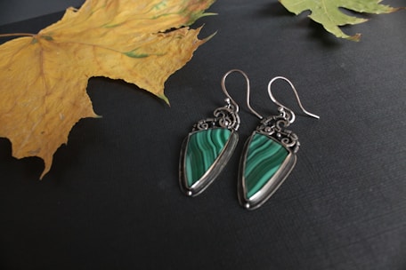 A pair of silver earrings with green malachite gemstones displayed on a dark surface with two dried autumn leaves placed nearby.