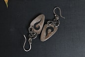 A pair of intricately designed metal earrings featuring an abstract swirl pattern. The earrings have a polished, silver-like finish and are set against a textured dark background. The intricate wirework and hook indicate careful craftsmanship.