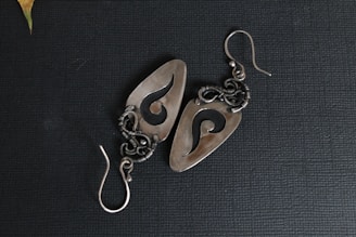 A pair of intricately designed metal earrings featuring an abstract swirl pattern. The earrings have a polished, silver-like finish and are set against a textured dark background. The intricate wirework and hook indicate careful craftsmanship.