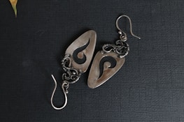 A pair of intricately designed metal earrings featuring an abstract swirl pattern. The earrings have a polished, silver-like finish and are set against a textured dark background. The intricate wirework and hook indicate careful craftsmanship.