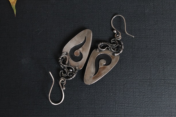 A pair of intricately designed metal earrings featuring an abstract swirl pattern. The earrings have a polished, silver-like finish and are set against a textured dark background. The intricate wirework and hook indicate careful craftsmanship.