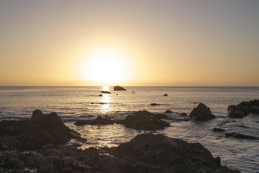 the sun is setting over the ocean with rocks in the foreground