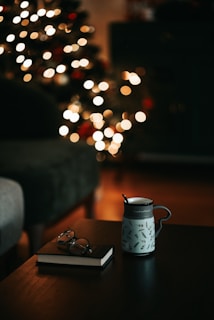A cozy indoor scene with a mug featuring a floral print and a spoon resting on a wooden table. Nearby, a closed book with a pair of glasses on top adds a studious touch. In the background, blurred festive lights suggest a decorated Christmas tree, creating a warm and inviting atmosphere.
