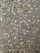A close-up view of a textured asphalt surface, with various small and medium-sized stones embedded within it. The surface appears slightly wet, enhancing the contrast and making the stones stand out. Some small twigs are also visible on the surface.