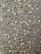 A close-up view of a textured asphalt surface, with various small and medium-sized stones embedded within it. The surface appears slightly wet, enhancing the contrast and making the stones stand out. Some small twigs are also visible on the surface.