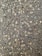 A close-up view of a textured asphalt surface, with various small and medium-sized stones embedded within it. The surface appears slightly wet, enhancing the contrast and making the stones stand out. Some small twigs are also visible on the surface.