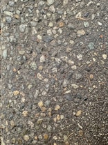A close-up view of a textured asphalt surface, with various small and medium-sized stones embedded within it. The surface appears slightly wet, enhancing the contrast and making the stones stand out. Some small twigs are also visible on the surface.