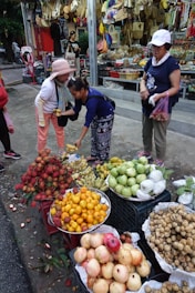 A bustling street market scene features a variety of colorful fruits displayed in baskets, including rambutans, bananas, oranges, pomelos, and more. Three women are engaged in a transaction; one is handing fruit to another while the third holds a bag. Behind them, there's a shop displaying woven bags, hats, and other goods.