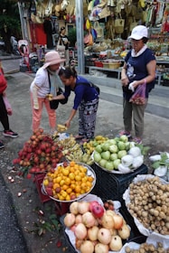 A bustling street market scene features a variety of colorful fruits displayed in baskets, including rambutans, bananas, oranges, pomelos, and more. Three women are engaged in a transaction; one is handing fruit to another while the third holds a bag. Behind them, there's a shop displaying woven bags, hats, and other goods.