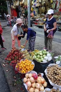 A bustling street market scene features a variety of colorful fruits displayed in baskets, including rambutans, bananas, oranges, pomelos, and more. Three women are engaged in a transaction; one is handing fruit to another while the third holds a bag. Behind them, there's a shop displaying woven bags, hats, and other goods.