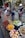 A bustling street market scene features a variety of colorful fruits displayed in baskets, including rambutans, bananas, oranges, pomelos, and more. Three women are engaged in a transaction; one is handing fruit to another while the third holds a bag. Behind them, there's a shop displaying woven bags, hats, and other goods.