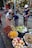 A bustling street market scene features a variety of colorful fruits displayed in baskets, including rambutans, bananas, oranges, pomelos, and more. Three women are engaged in a transaction; one is handing fruit to another while the third holds a bag. Behind them, there's a shop displaying woven bags, hats, and other goods.