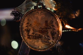 A close-up of a decorative Christmas ornament hanging on a tree. The ornament is round, adorned with intricate golden detailing along its edges. Inside the ornament, there is a wintery scene featuring snow-covered trees, a house, and a tall tower. The ornament is topped with a translucent ribbon tied into a bow, adding an elegant touch. Warm white lights and dark green pine needles surround the ornament, contributing to a festive atmosphere.