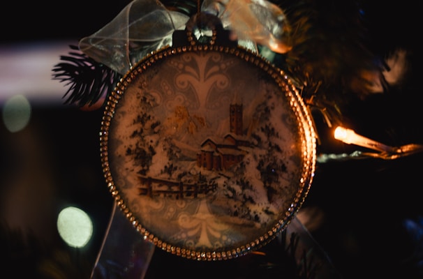 A close-up of a decorative Christmas ornament hanging on a tree. The ornament is round, adorned with intricate golden detailing along its edges. Inside the ornament, there is a wintery scene featuring snow-covered trees, a house, and a tall tower. The ornament is topped with a translucent ribbon tied into a bow, adding an elegant touch. Warm white lights and dark green pine needles surround the ornament, contributing to a festive atmosphere.