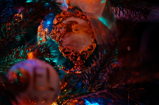 A close-up of a decorated Christmas tree features ornate ornaments. The image displays rich details with an ornament that has an intricate golden design and a vintage image of a child. The background includes a blurred ornament with the letters 'EL' visible and pine needles illuminated by colorful lights.