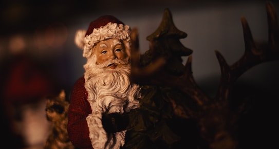 A figurine of Santa Claus is shown, featuring his traditional red outfit and white beard. He is standing next to a reindeer statue with antlers. The background is blurred, emphasizing the focus on the figurines.