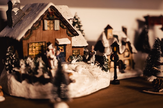 A charming winter village scene with miniature snow-covered houses and figurines creating a cozy, nostalgic atmosphere. Illuminated windows suggest warmth inside, while a lamppost adds to the quaint setting.