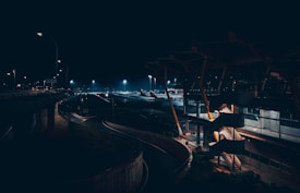 An airport scene at night featuring dimly lit terminals and a runway. The area is illuminated by streetlights, creating a contrast with the dark sky. Airplanes are visible in the background, parked at the gate. Spiral staircases and modern architectural elements add to the futuristic vibe.