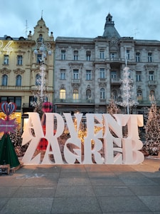 Large white letters spell out 'Advent Zagreb' in front of a backdrop of ornate historic buildings. The scene is decorated with Christmas trees adorned with lights, giving a festive atmosphere. Decorative elements include colorful ornaments and stylized trees, while the sky is overcast, contributing to the wintery feel.