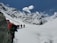 A group of hikers is trekking through a snowy mountainous landscape under a clear blue sky. The snow-covered mountains loom majestically in the background, while the hikers wear colorful gear and carry backpacks. The scene suggests a challenging and adventurous journey through pristine, untouched nature.