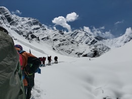 A group of hikers is trekking through a snowy mountainous landscape under a clear blue sky. The snow-covered mountains loom majestically in the background, while the hikers wear colorful gear and carry backpacks. The scene suggests a challenging and adventurous journey through pristine, untouched nature.