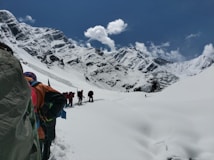A group of hikers is trekking through a snowy mountainous landscape under a clear blue sky. The snow-covered mountains loom majestically in the background, while the hikers wear colorful gear and carry backpacks. The scene suggests a challenging and adventurous journey through pristine, untouched nature.