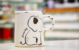A ceramic mug with an elephant design, featuring a stylized drawing of a cute elephant and the words 'dream big'. The mug has a glossy finish and includes decorative heart patterns on the handle. In the background, there is a blurred image of shelves with various items.
