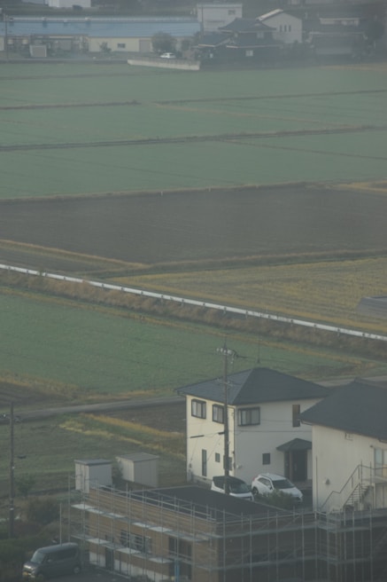 Farmland with rectangular fields in shades of green and brown, interspersed with pathways. In the foreground, there are residential buildings with cars parked next to them. A power line and pole can also be seen, along with scaffolding around one of the buildings.