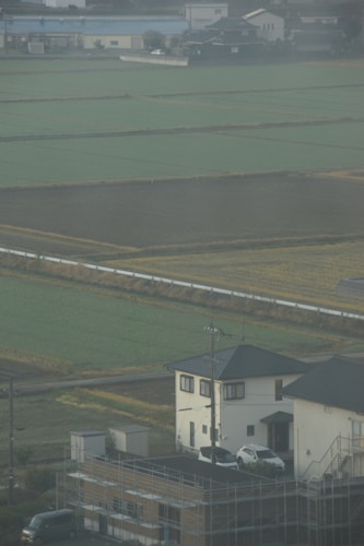 Farmland with rectangular fields in shades of green and brown, interspersed with pathways. In the foreground, there are residential buildings with cars parked next to them. A power line and pole can also be seen, along with scaffolding around one of the buildings.