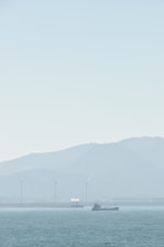A serene seascape with a ship sailing on calm waters. In the background, several wind turbines stand against a hazy mountainous backdrop, and a small white plume of smoke or steam rises from a structure.