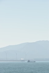 A serene seascape with a ship sailing on calm waters. In the background, several wind turbines stand against a hazy mountainous backdrop, and a small white plume of smoke or steam rises from a structure.