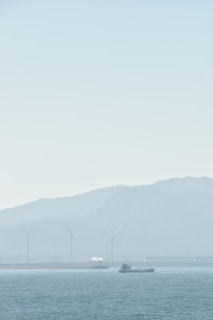 A serene seascape with a ship sailing on calm waters. In the background, several wind turbines stand against a hazy mountainous backdrop, and a small white plume of smoke or steam rises from a structure.