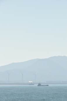 A serene seascape with a ship sailing on calm waters. In the background, several wind turbines stand against a hazy mountainous backdrop, and a small white plume of smoke or steam rises from a structure.