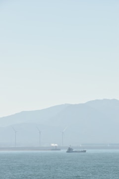 A serene seascape with a ship sailing on calm waters. In the background, several wind turbines stand against a hazy mountainous backdrop, and a small white plume of smoke or steam rises from a structure.